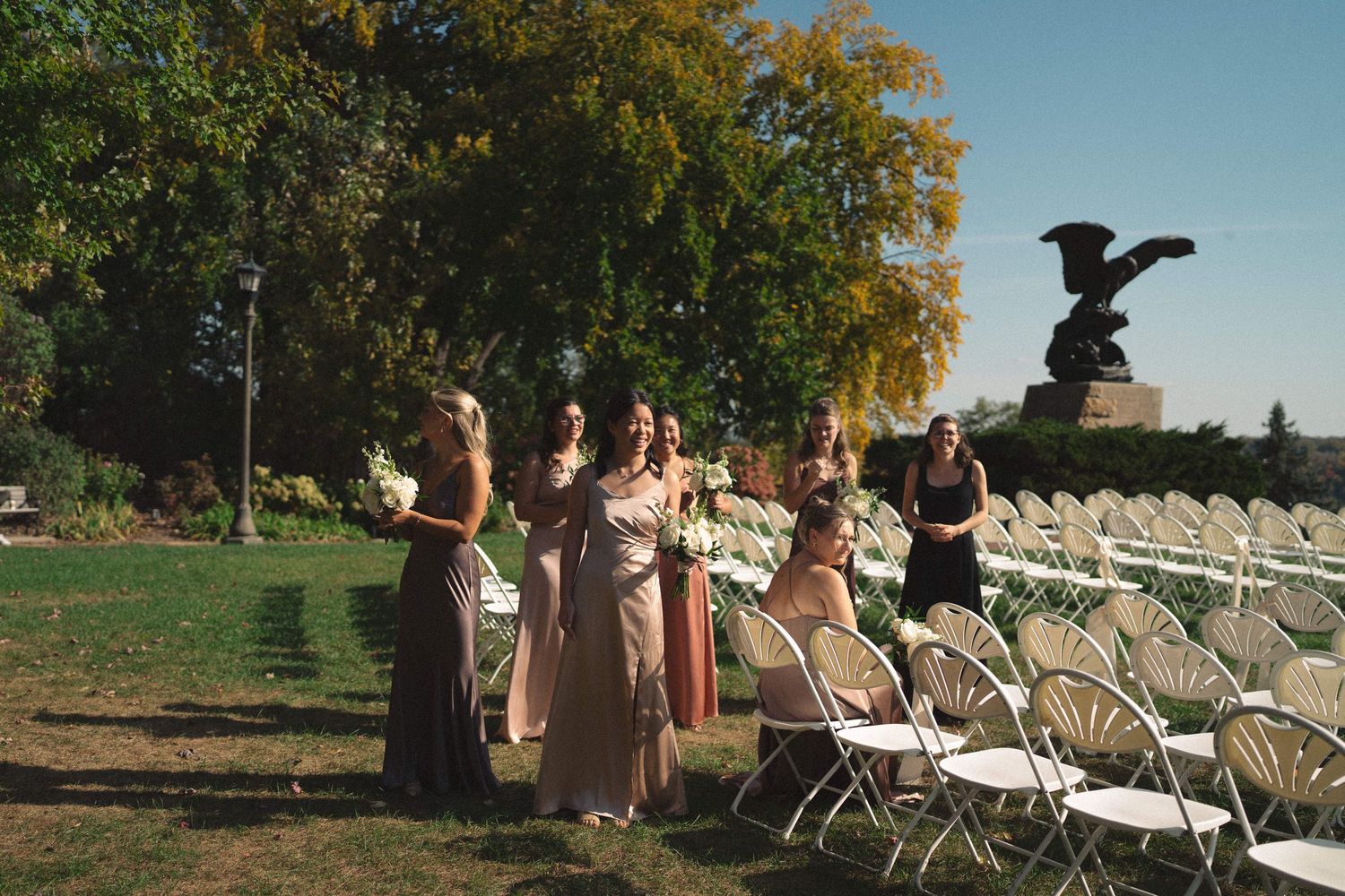 Wedding party members stand near white chairs with an eagle statue in the background.