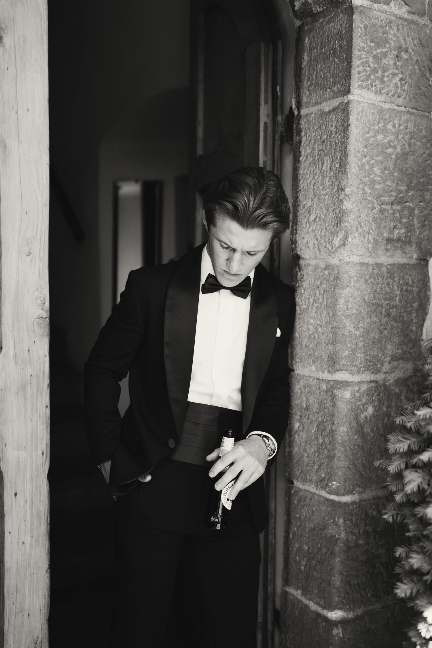 Black and white photo of an elegant young man in a tuxedo with bow tie, leaning against a stone pillar.