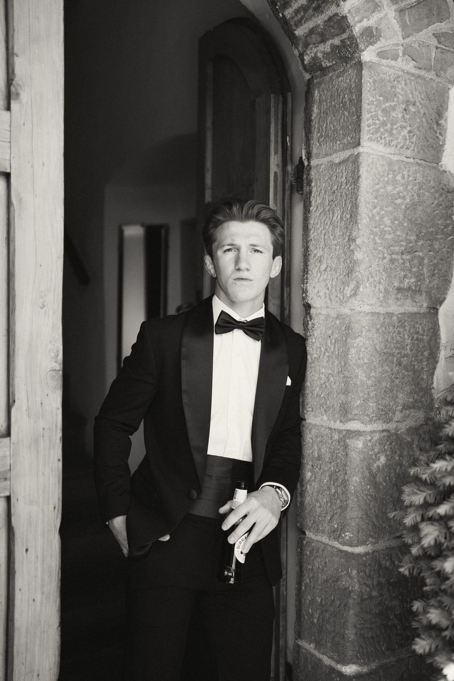 Young man in black tuxedo and bow tie leaning against stone archway in elegant black and white portrait photo.