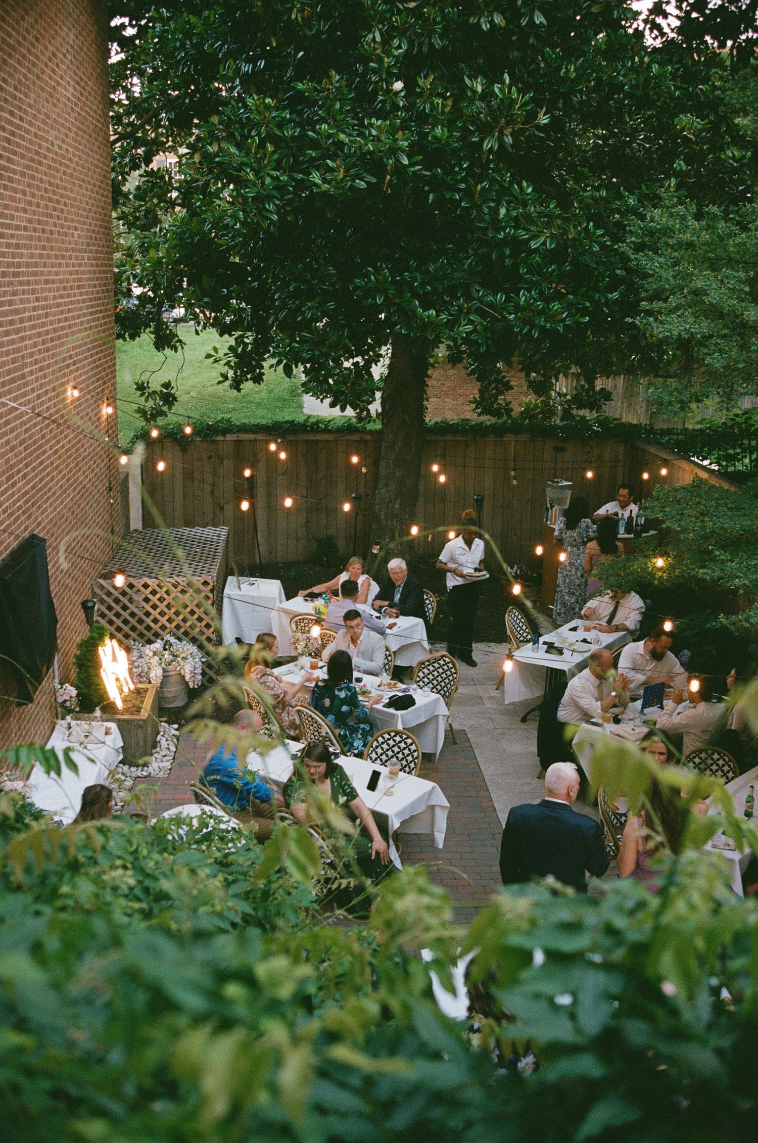 An outdoor wedding reception with string lights and guests dining on a patio surrounded by greenery.
