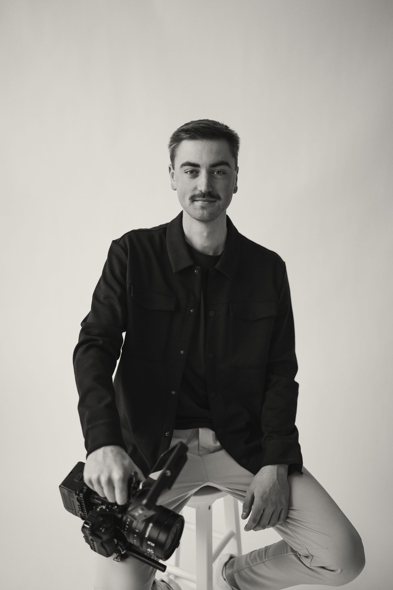 Professional photographer in black jacket poses with camera in studio setting.