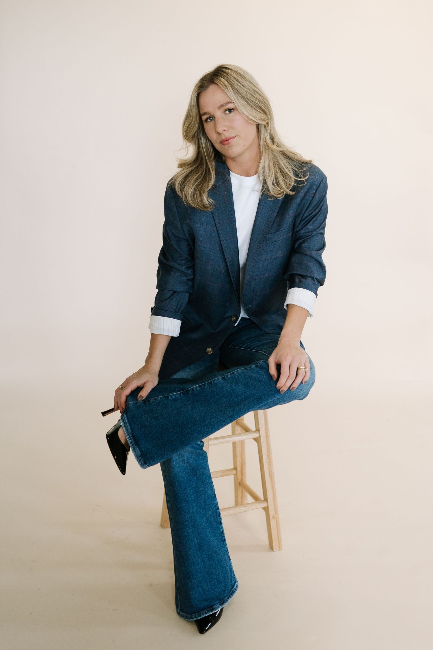 Professional seated pose in navy blazer and flared jeans on wooden stool.