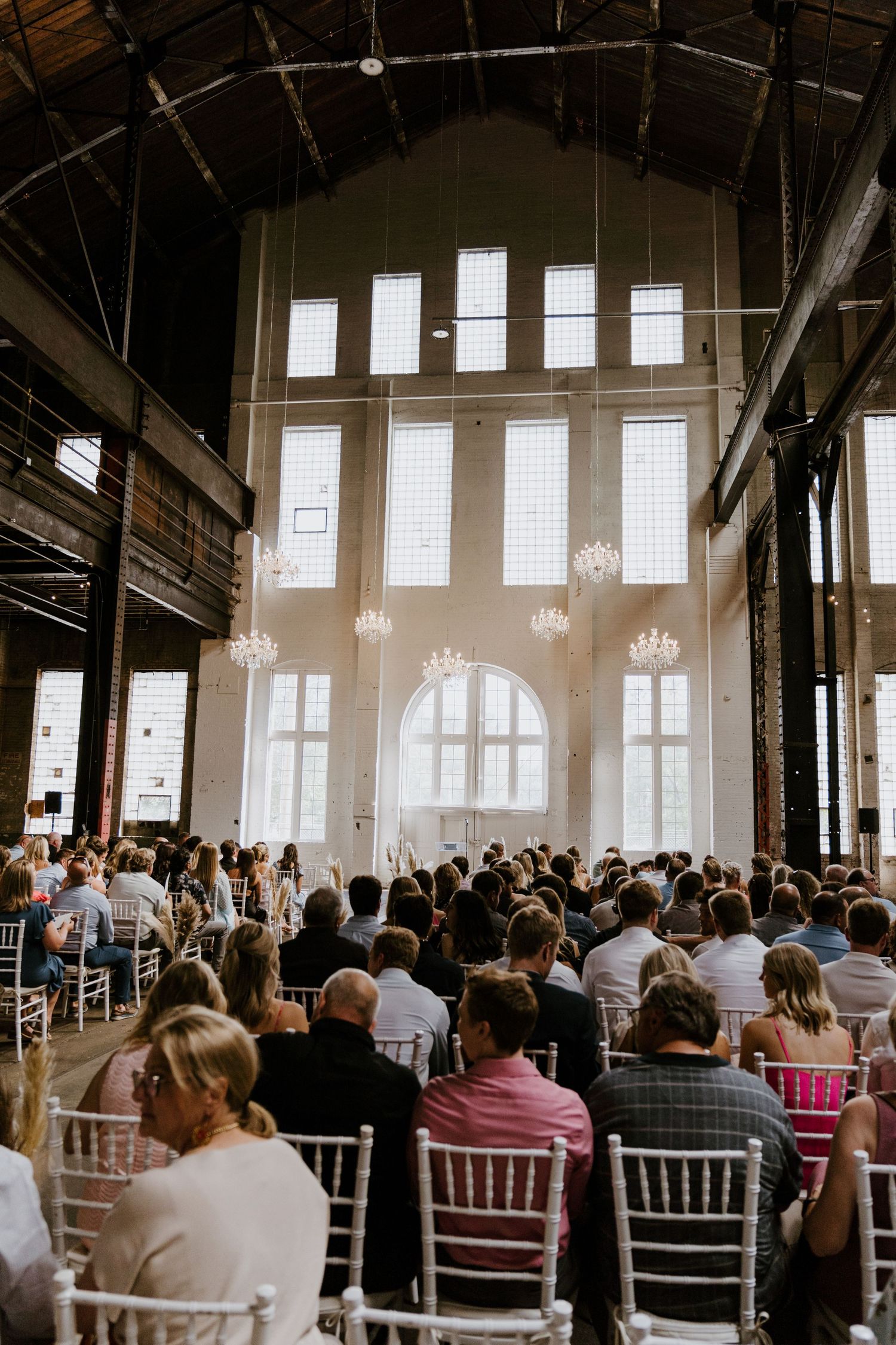 Wedding ceremony venue with rows of chairs and hanging chandeliers in an elegant industrial-style space.