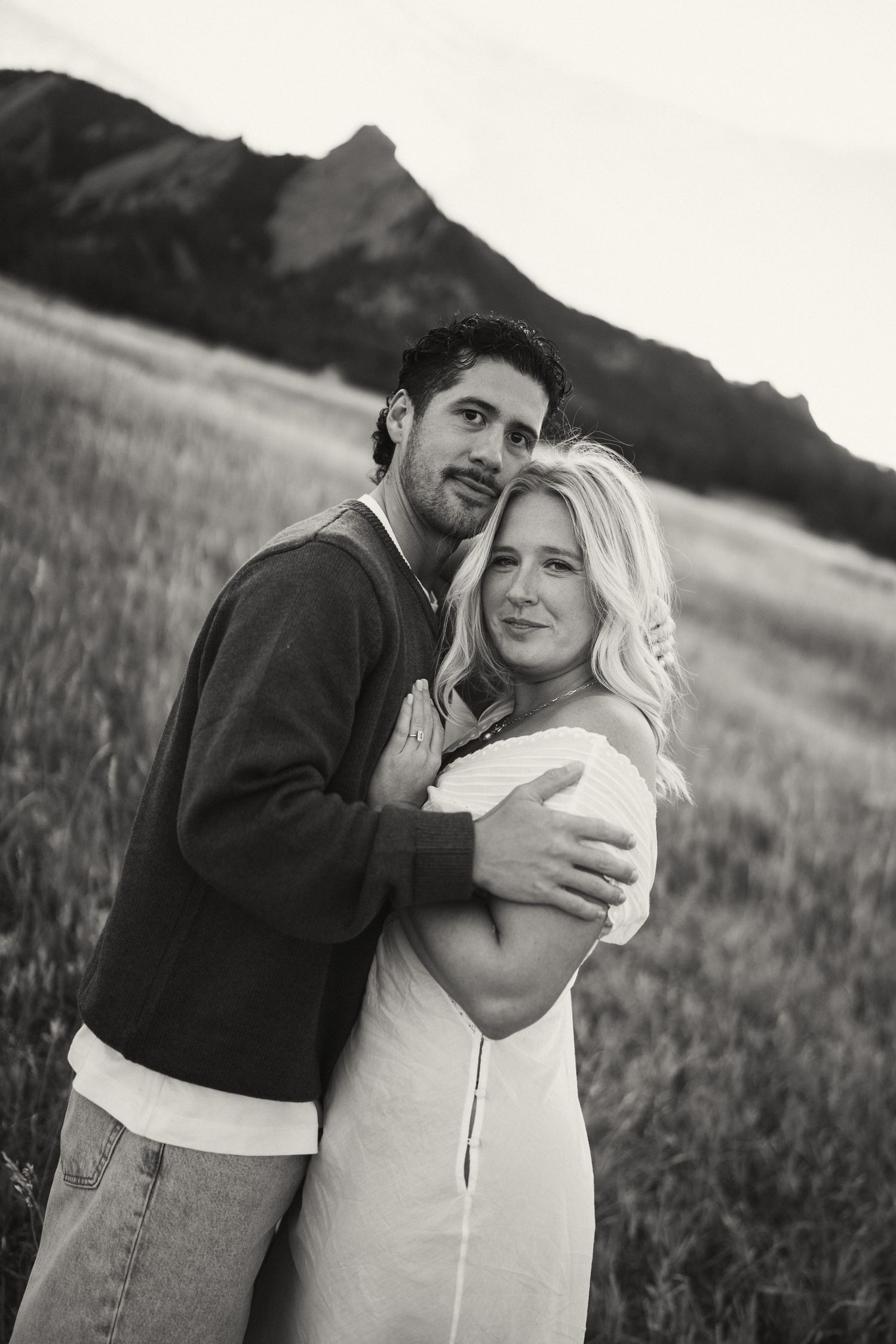 Black and white portrait of a couple embracing in a field with mountains behind them.
