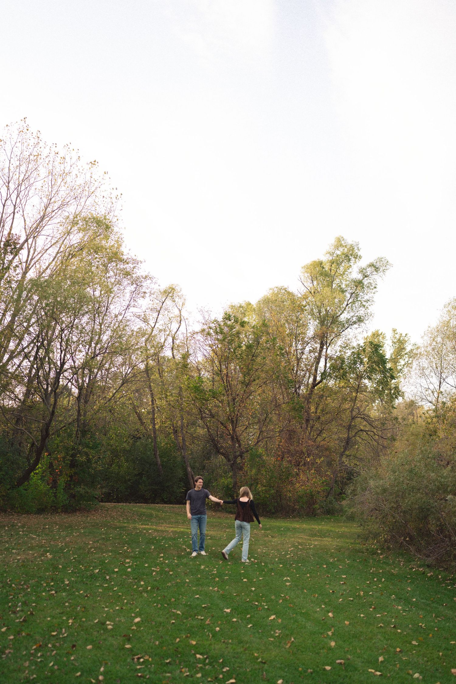 Two people walking and interacting in a spacious green field surrounded by trees.
