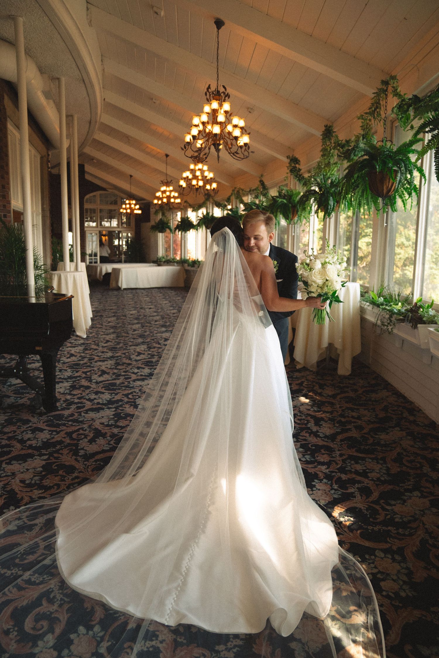 Long flowing white wedding dress photographed in an elegant venue with chandeliers and greenery.