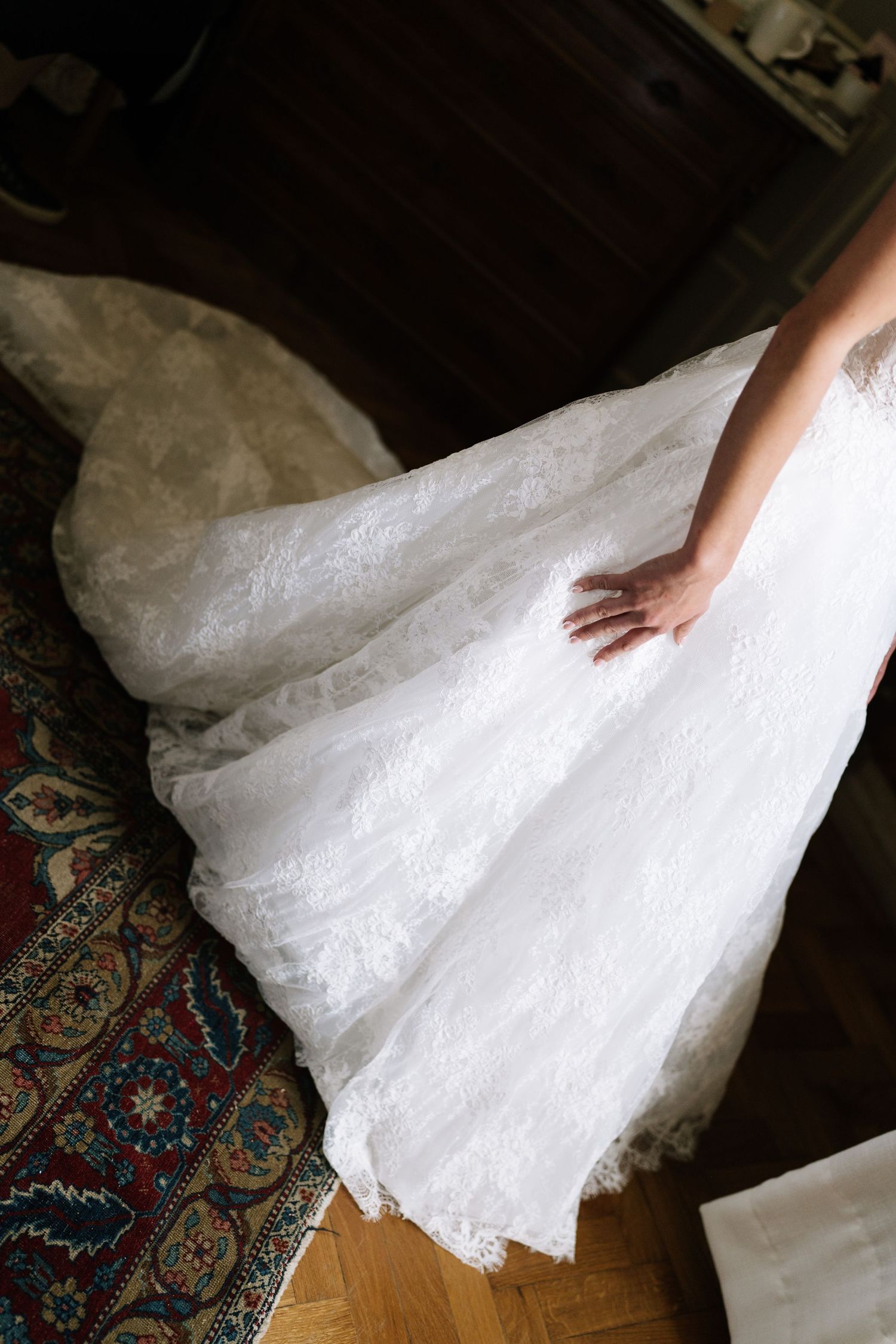 Bride in a white lace wedding gown with a flowing train, gently touching the skirt on a wooden floor.