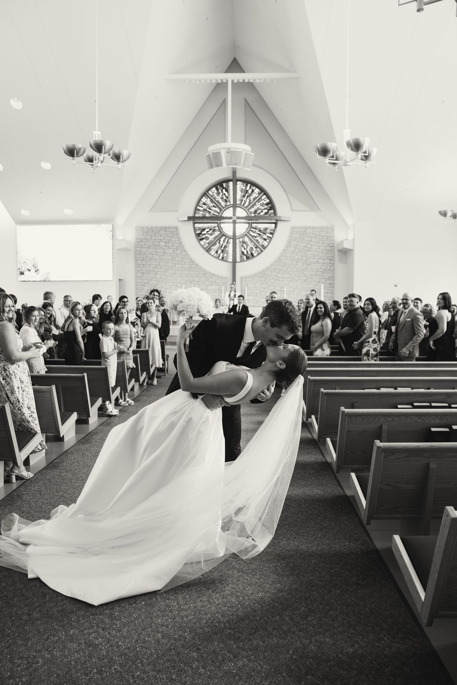 Dramatic wedding kiss in a church with a large cross and stained glass window in the background.