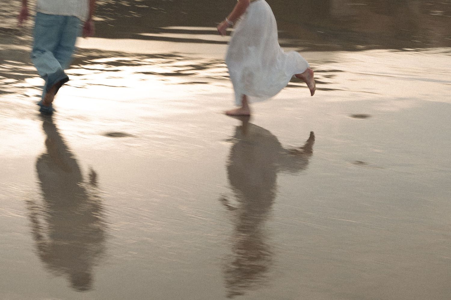 Two people running barefoot on wet sand at sunset, their reflections shimmering in the shallow water below.
