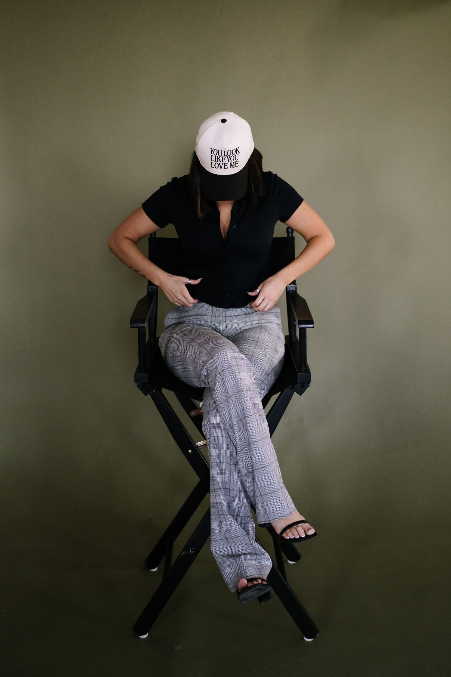 Person in black top and plaid pants sitting in director's chair with white cap.