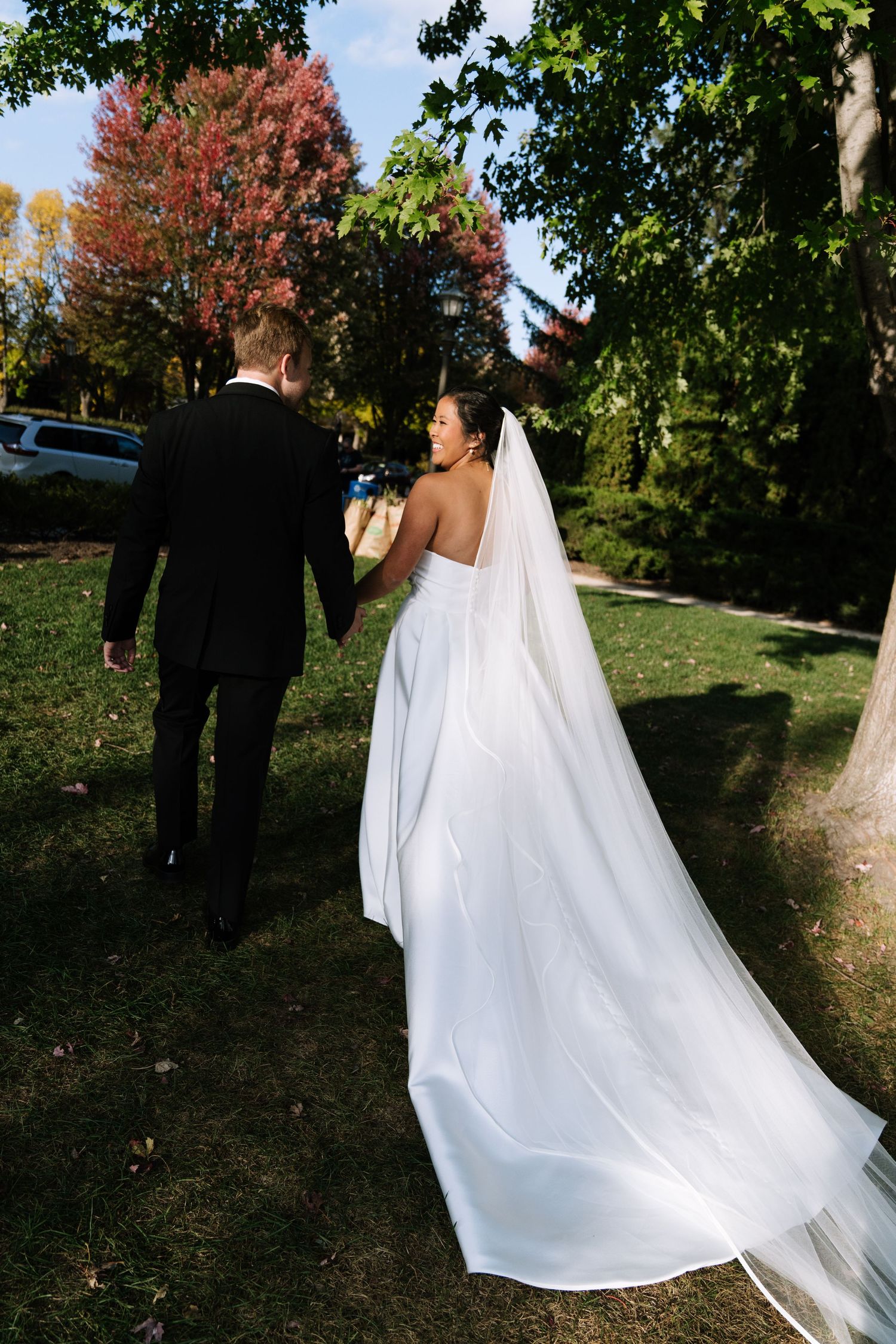 Bride in flowing white dress walks with groom through autumn trees.
