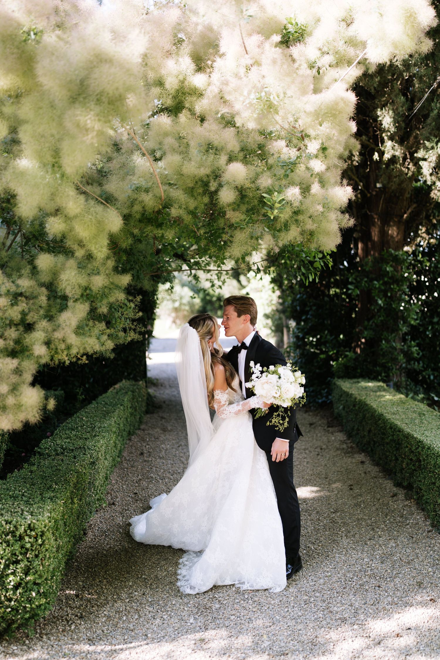 Bride and groom share a romantic kiss under lush green canopy in a formal garden pathway.