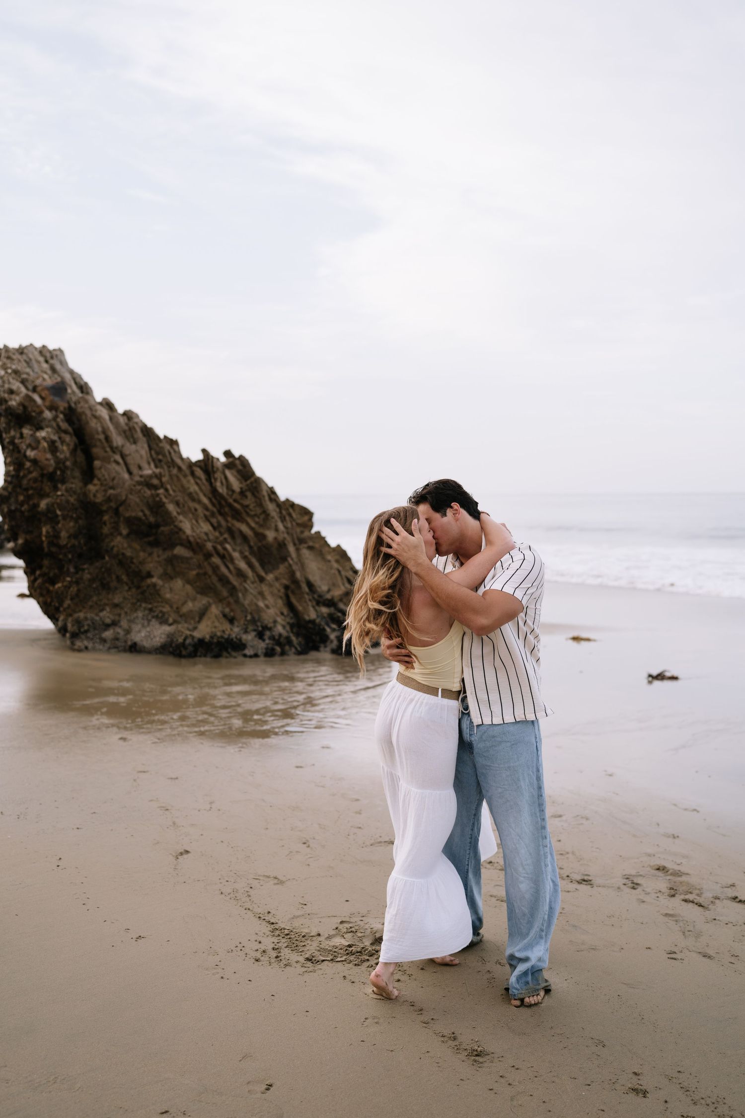 A couple shares a romantic kiss on a sandy beach with rocky cliffs and ocean waves in the background.