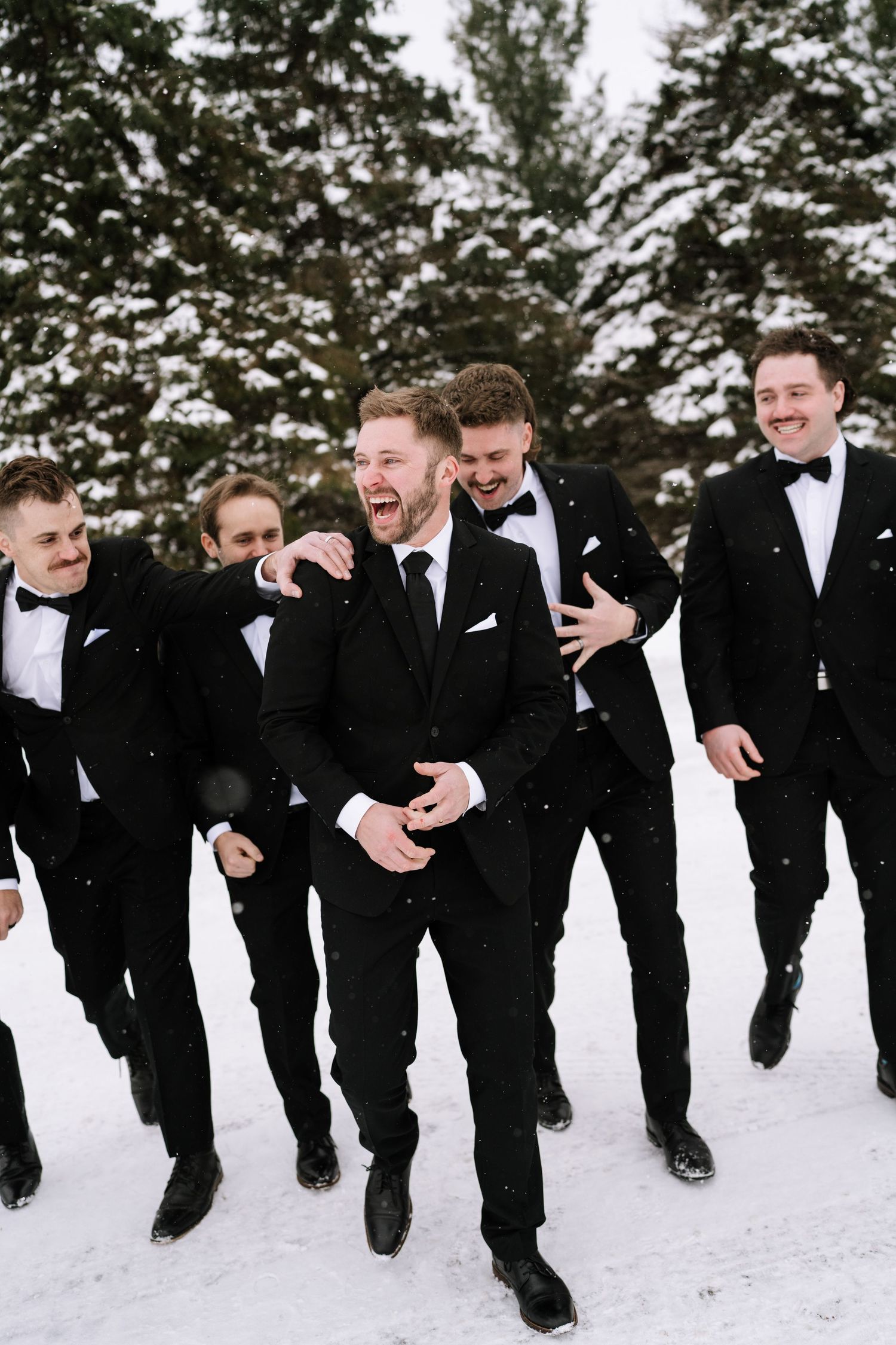 Groomsmen in black tuxedos running and jumping in snow during winter wedding photoshoot.