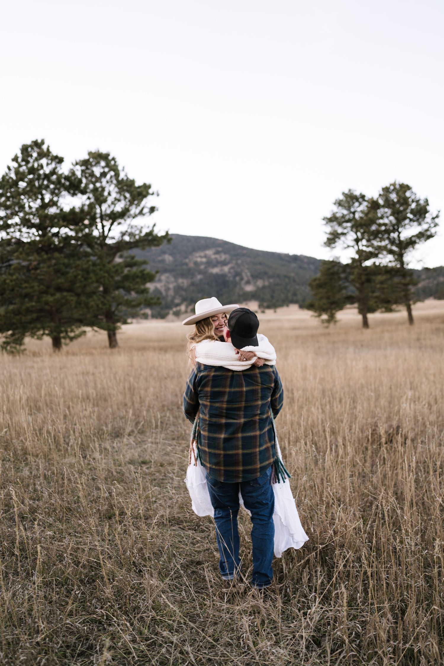A couple embraces in a rustic field with mountains and pine trees in the background.