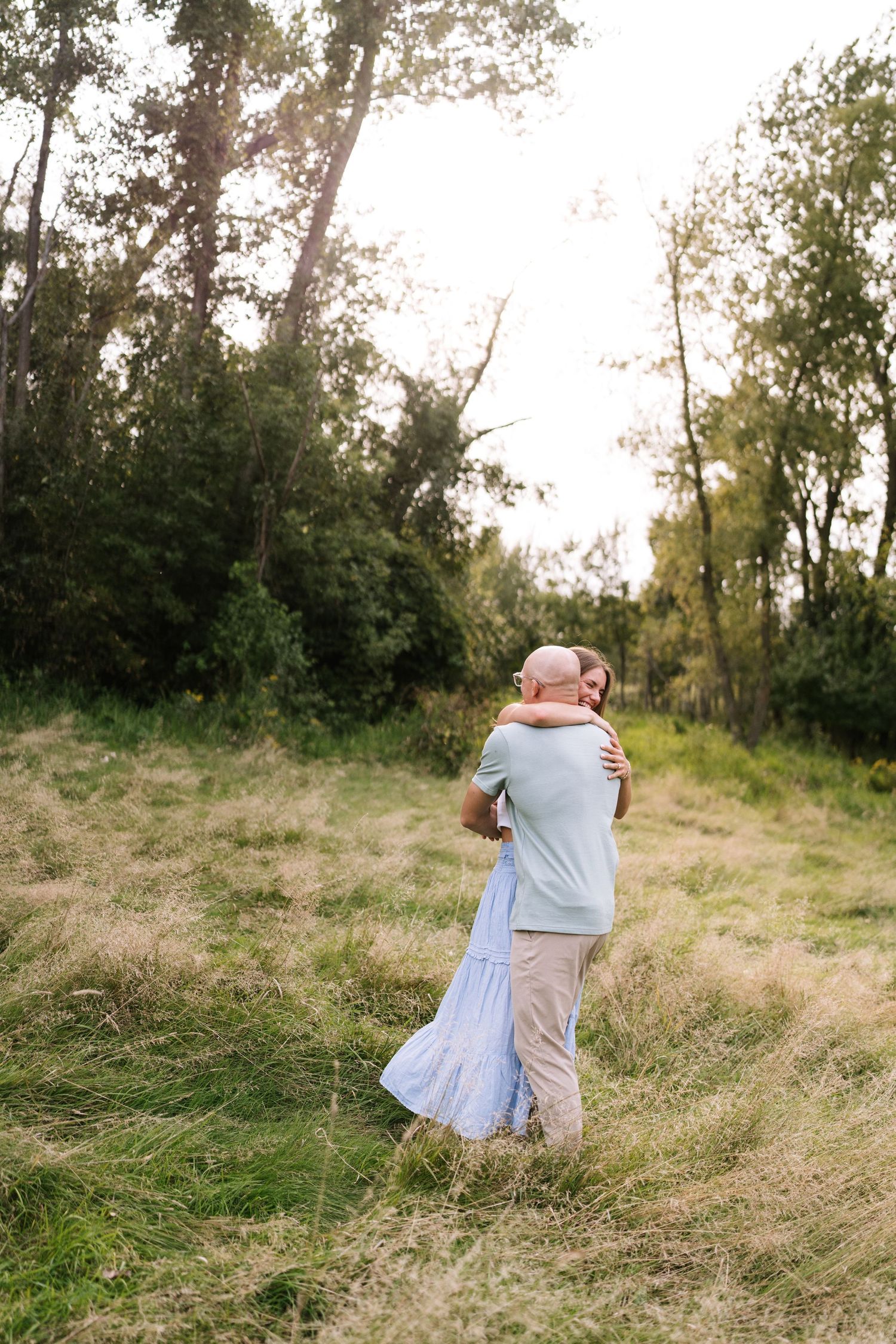 A couple shares an intimate embrace in a sunlit meadow surrounded by trees.