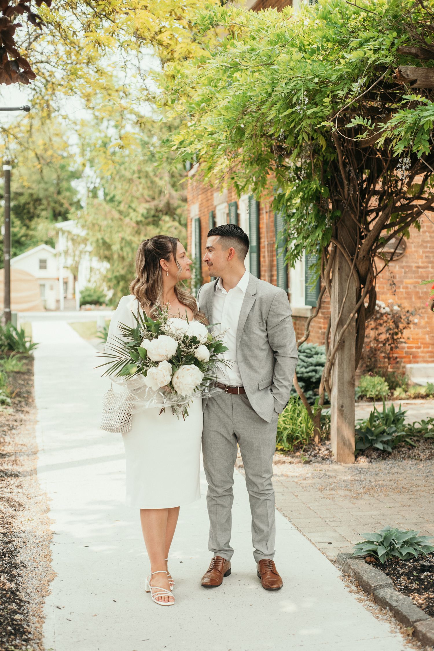 Wedding portrait of a couple walking hand in hand outdoors, smiling at each other beneath greenery with a bouquet and elegant attire.