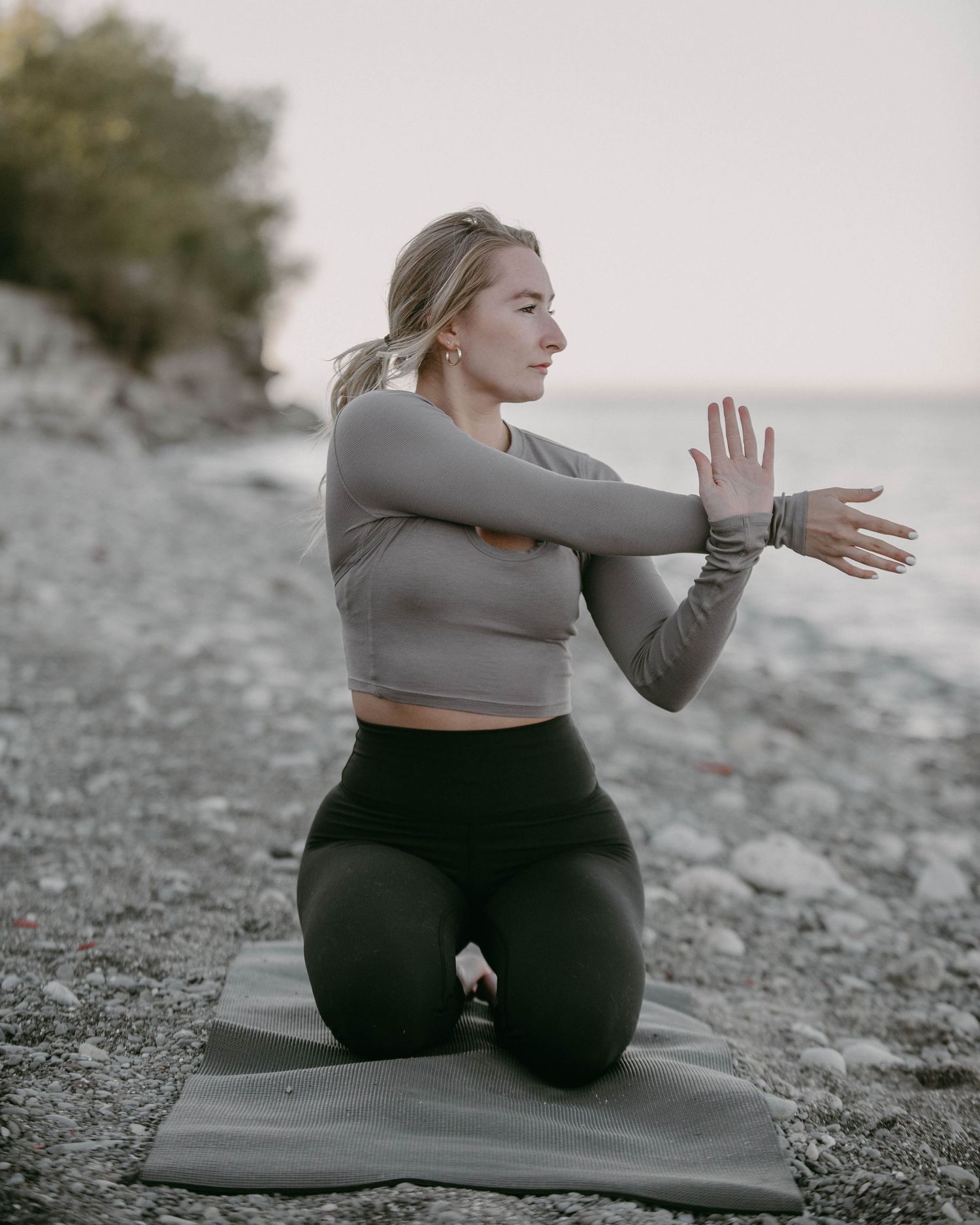 Lifestyle portrait of a woman stretching on a yoga mat by the water, wearing athletic wear and practicing mindful movement outdoors.