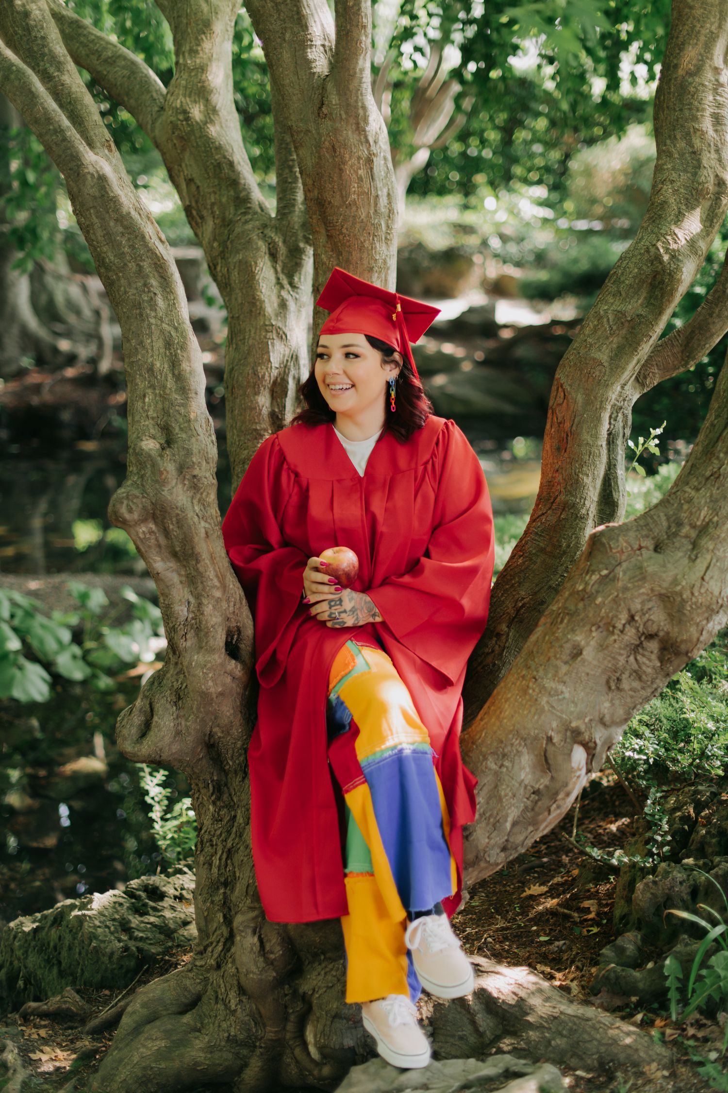Outdoor graduation portrait of a graduate in a red cap and gown sitting in a tree, smiling and holding an apple in a natural setting.