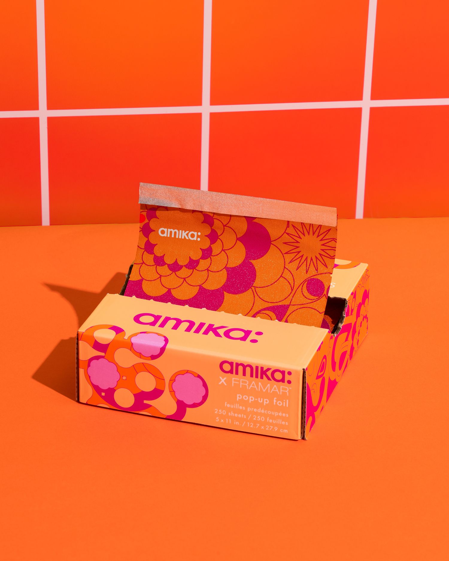 Studio product photography of Amika x Framar pop-up foil packaging styled on an orange background with bold, colorful branding.