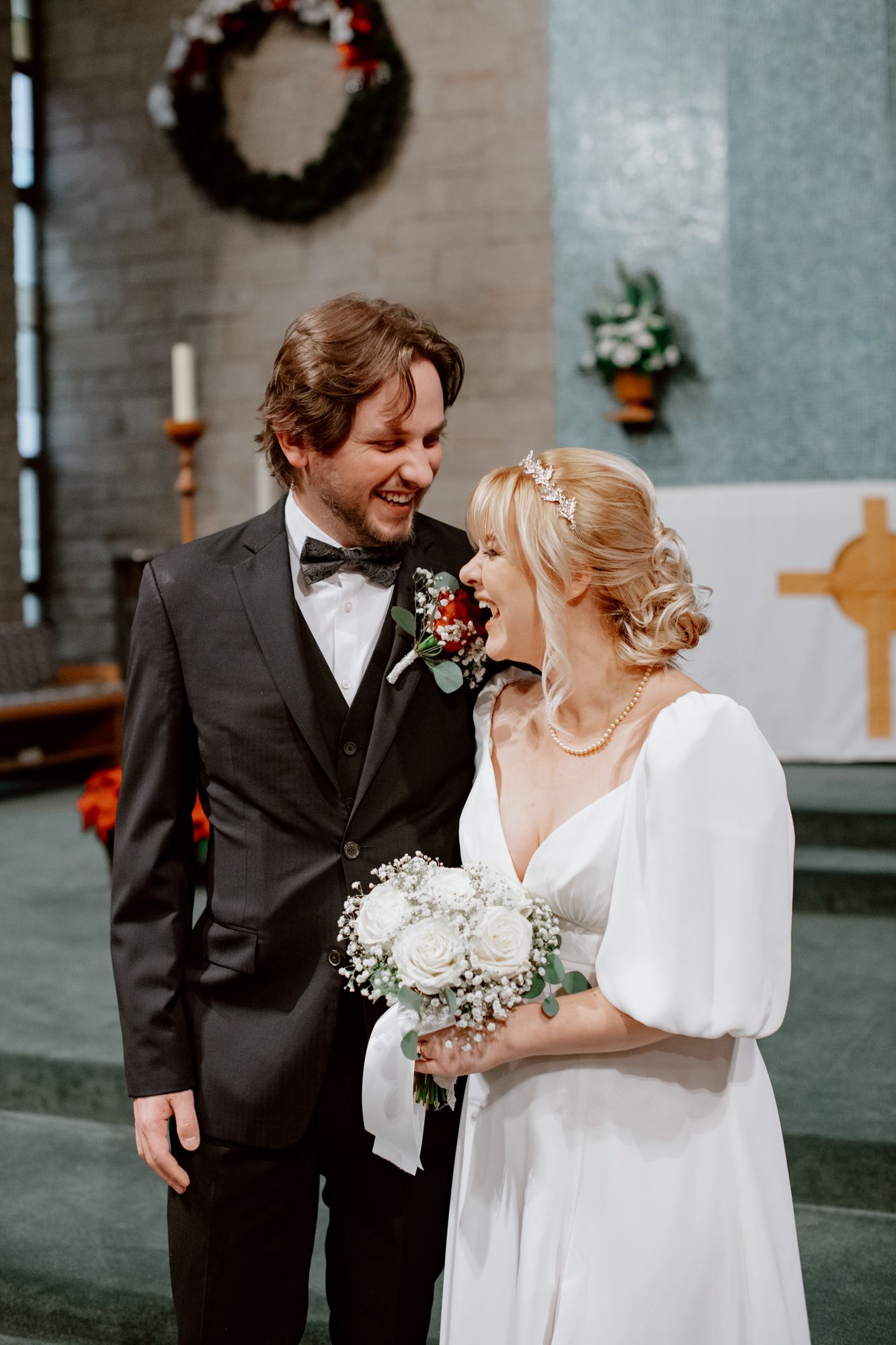 Candid wedding portrait of a bride and groom laughing together indoors, holding a bouquet during a joyful ceremony moment.