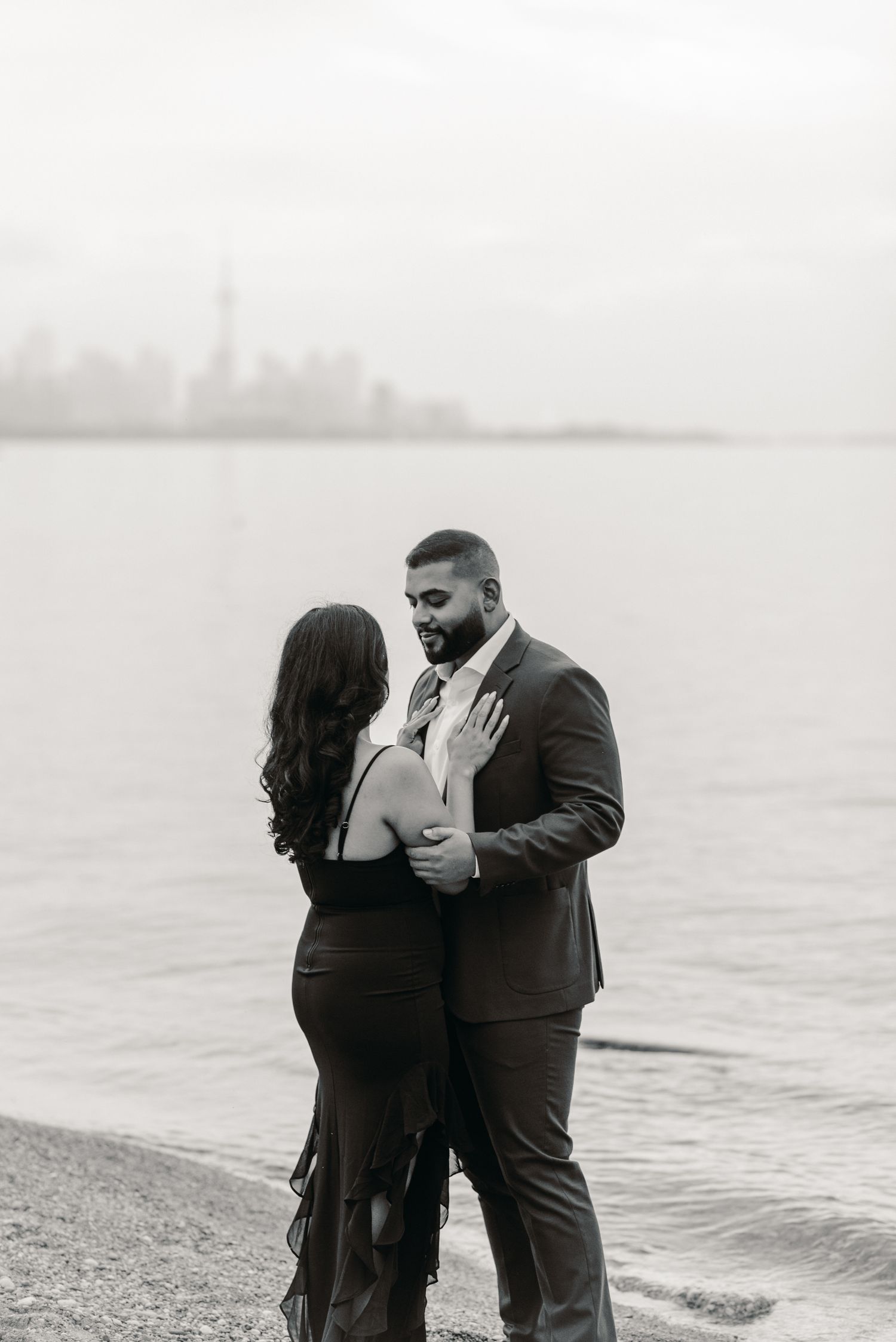 Black and white engagement portrait of a couple embracing by the water, sharing an intimate moment during a proposal photoshoot.