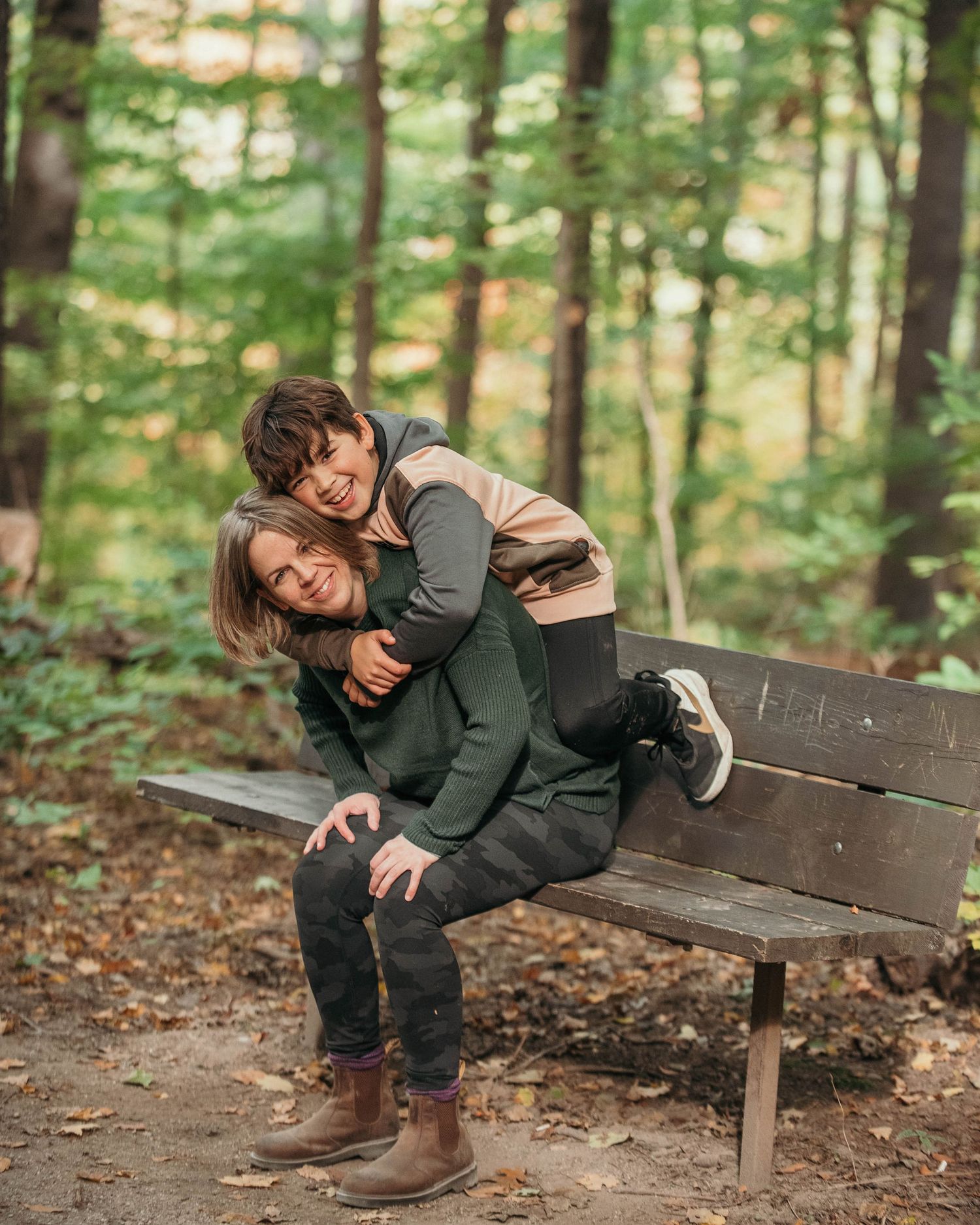 Lifestyle family portrait of a parent and child laughing on a park bench in a wooded setting during a candid outdoor moment.