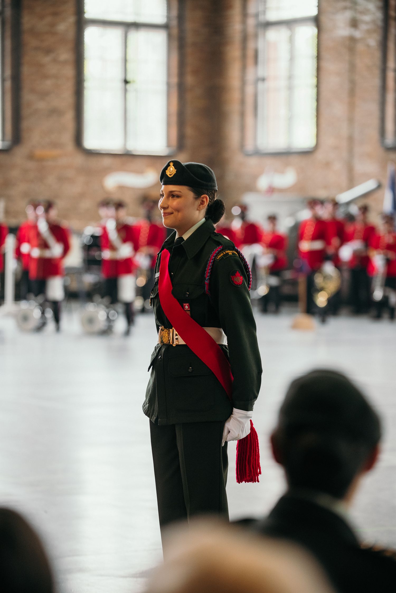 Ceremonial portrait of a uniformed service member standing proudly indoors during a formal military event with a band in the background.