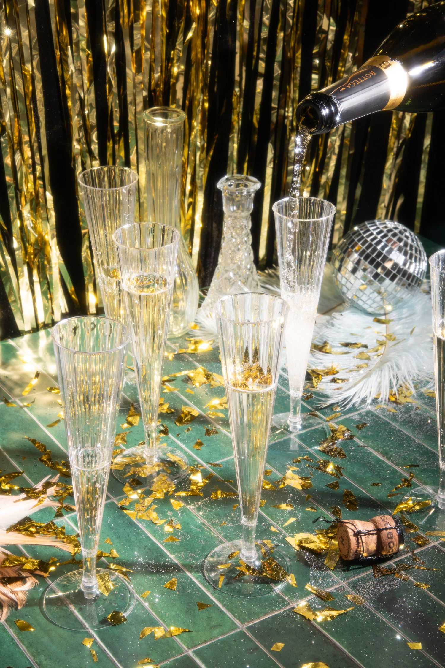 Lifestyle product photo of champagne being poured into crystal flutes with gold confetti, disco ball accents, and a celebratory party setup.