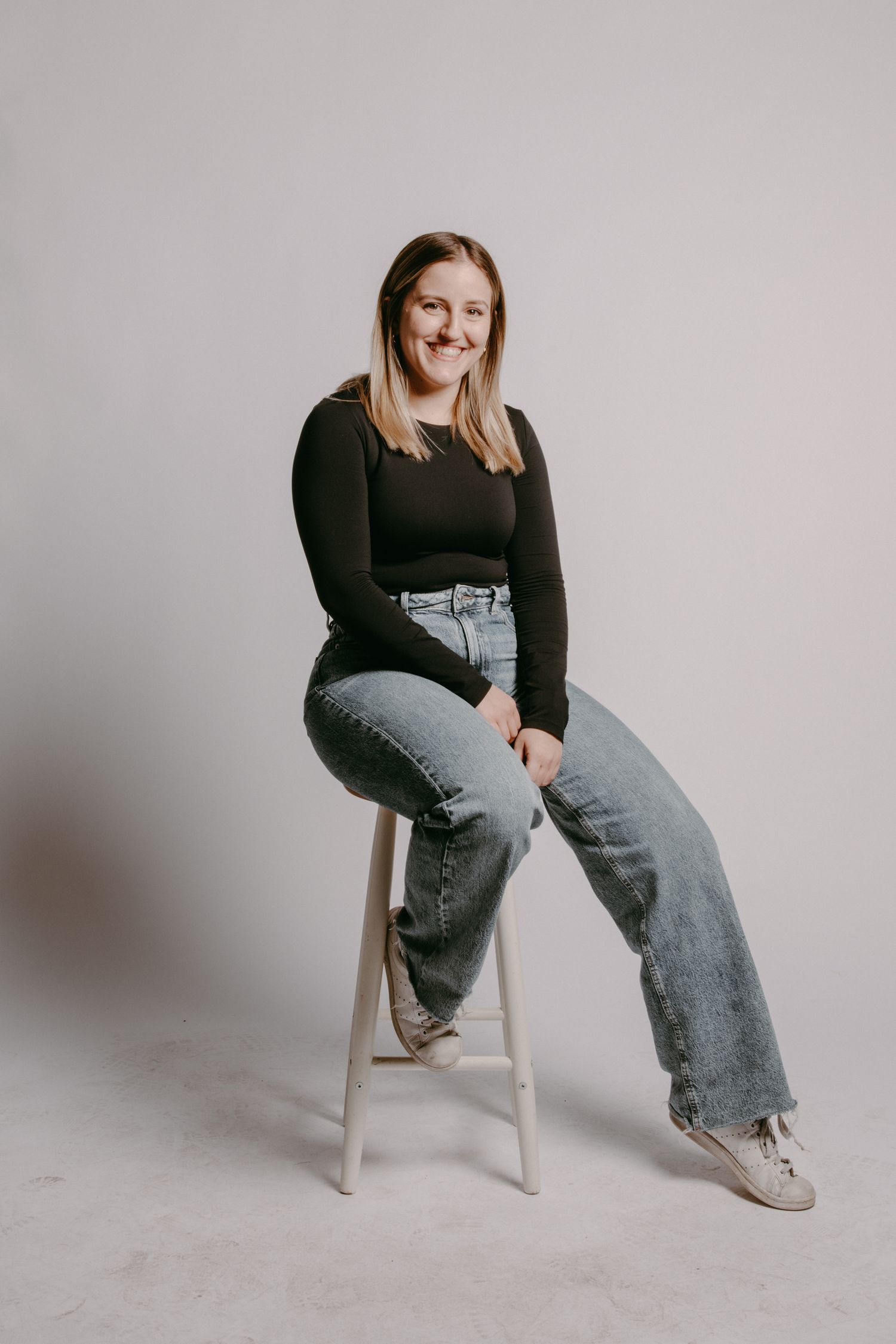 Studio portrait of Niagara Falls–based photographer Megan W sitting on a stool, smiling, wearing a black top, blue jeans, and white sneakers