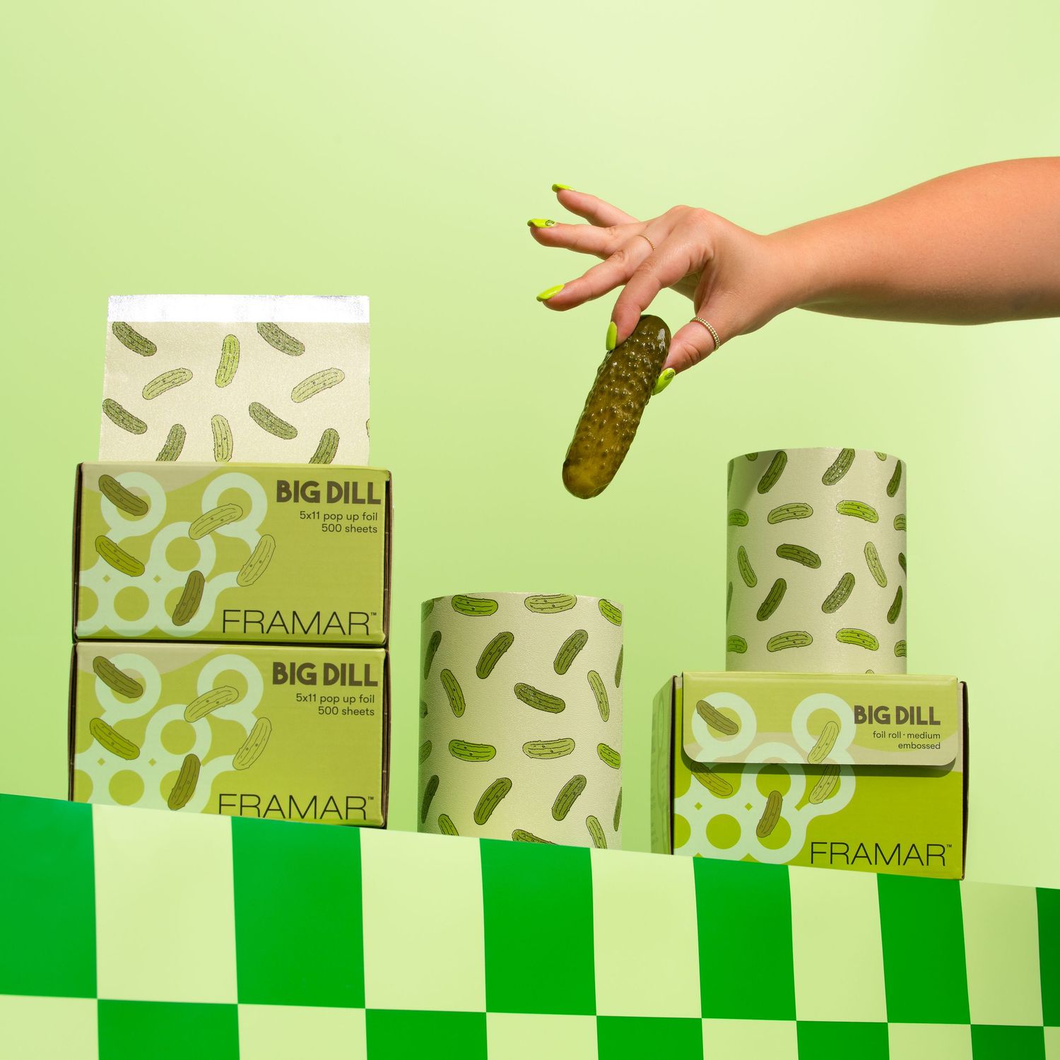 Studio product photography of Framar Big Dill hair foil packaging styled with green tones, pickle graphics, and a playful hand holding a pic