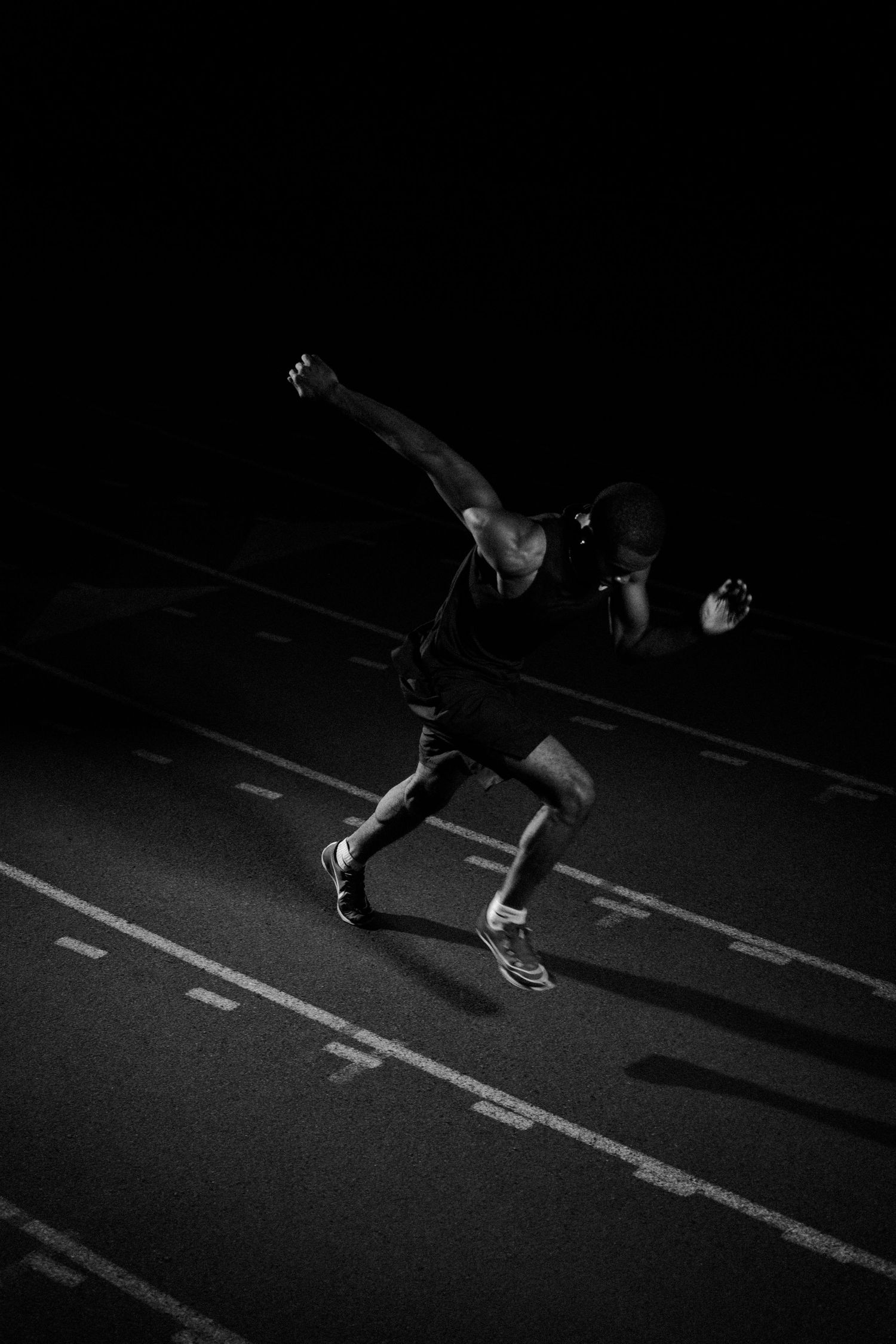 Black and white photo of an athlete sprinting on a track at night, captured mid-stride with dynamic motion.
