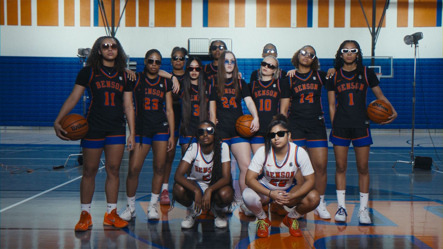 Basketball team poses in orange jerseys on gym court, two players kneel in front wearing sunglasses.