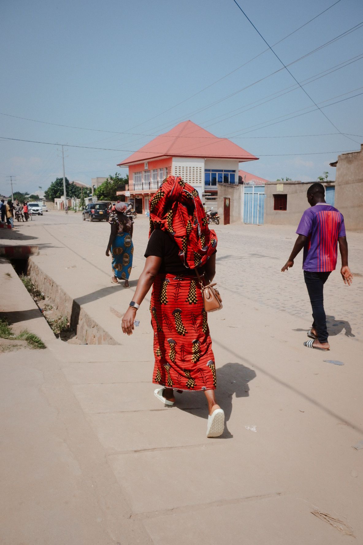 People walking along a dusty street with small buildings and red roofs visible.