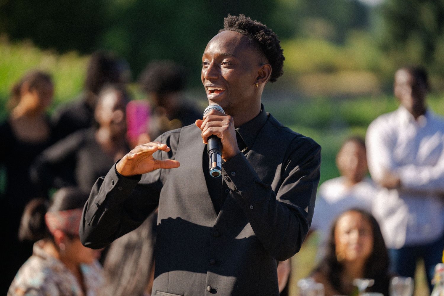 A young person speaks into a microphone outdoors at a gathering, wearing a black blazer with a crowd behind them.