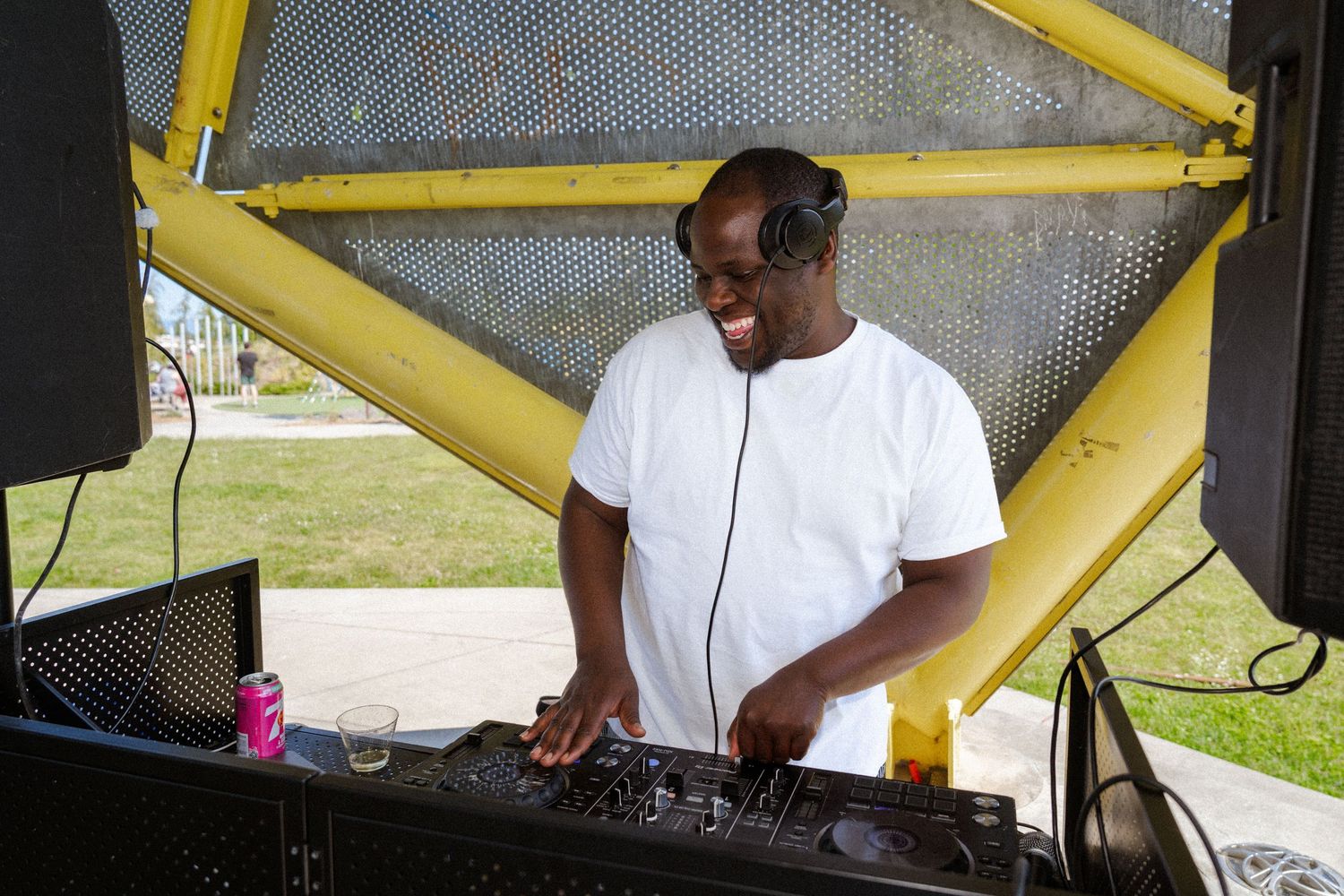 A smiling DJ wearing headphones and a white t-shirt mixing music outdoors at a sunny event.