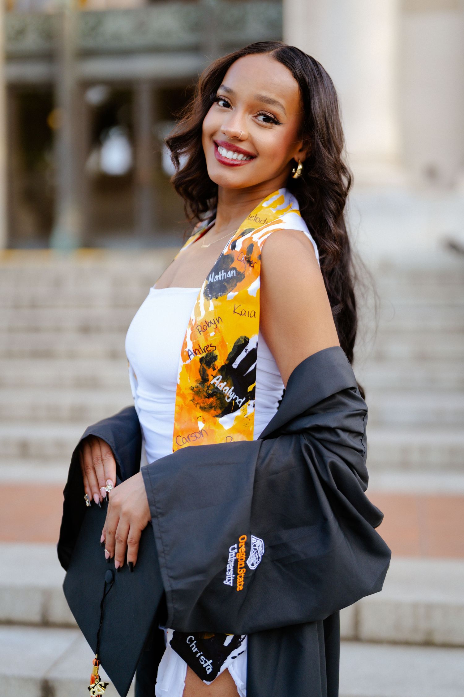 Smiling young woman in graduation gown and colorful sash poses on steps, holding cap.
