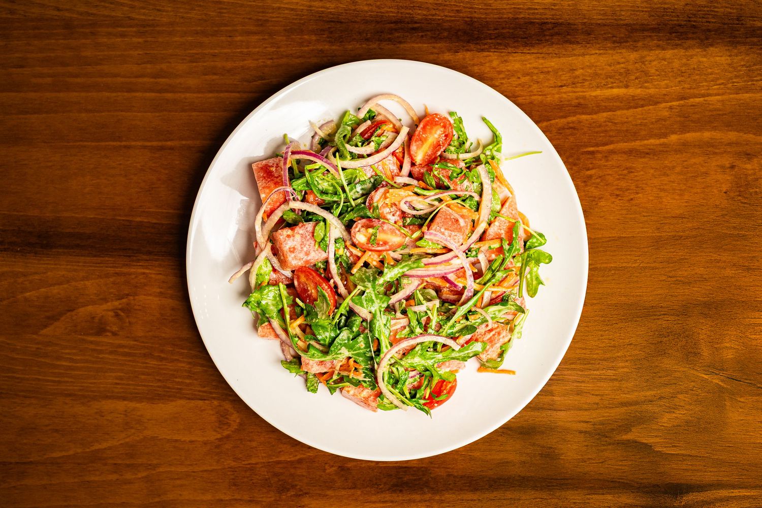 Fresh arugula salad with tomatoes and red onion on a white plate atop a wooden table.