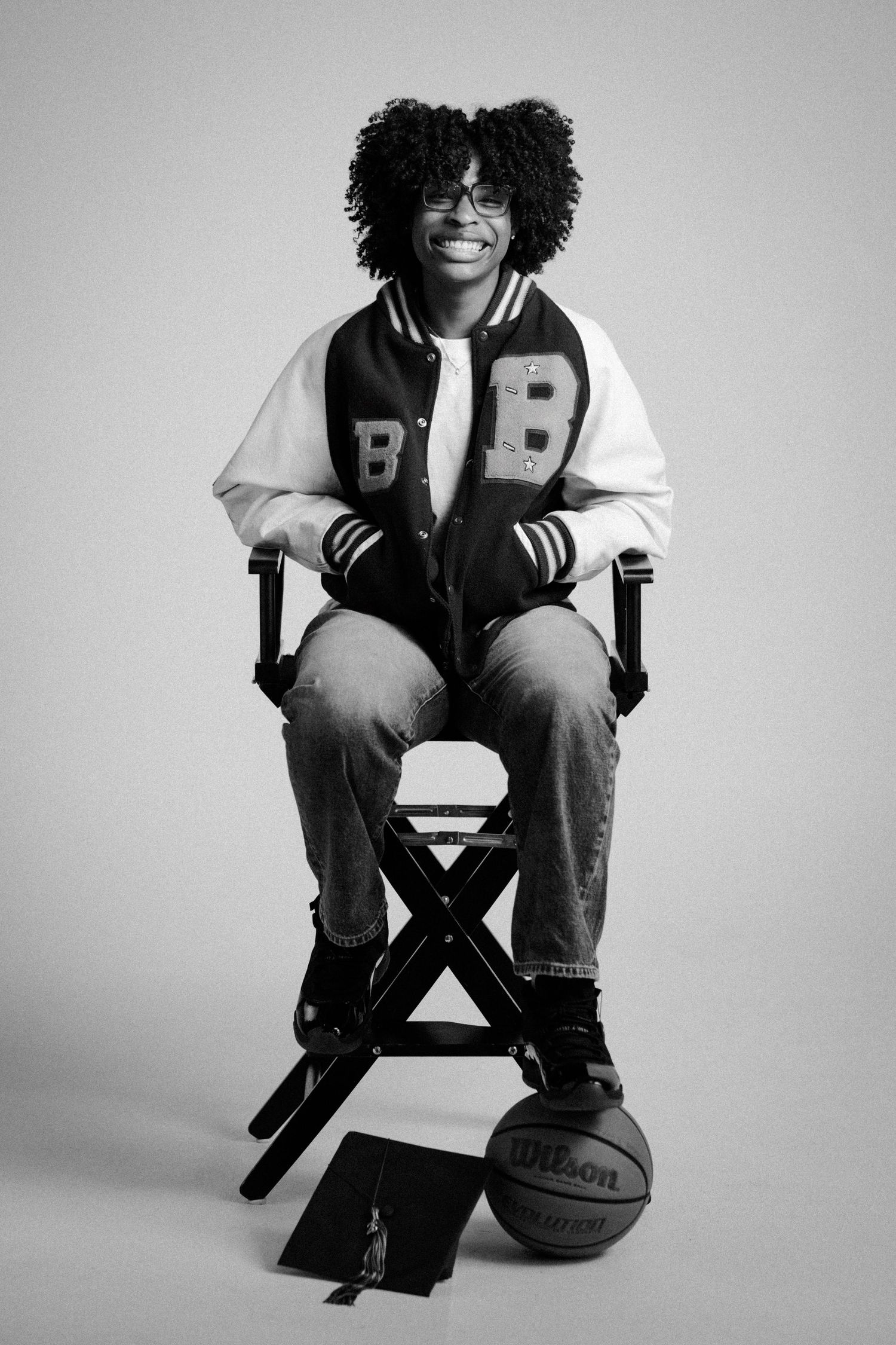 Young person in varsity jacket sitting on director's chair with Wilson basketball, black and white portrait photo.
