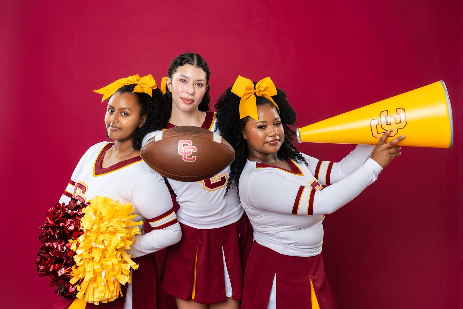 Cheerleaders in burgundy and white uniforms pose with pom poms, football and megaphone against red background.