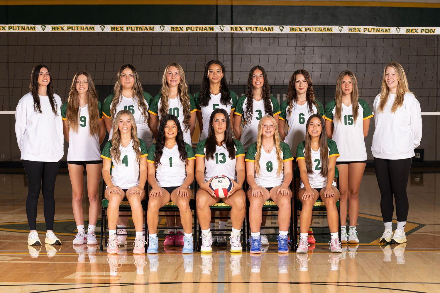 High school volleyball team poses for official team photo wearing white uniforms in front of black gymnasium wall.