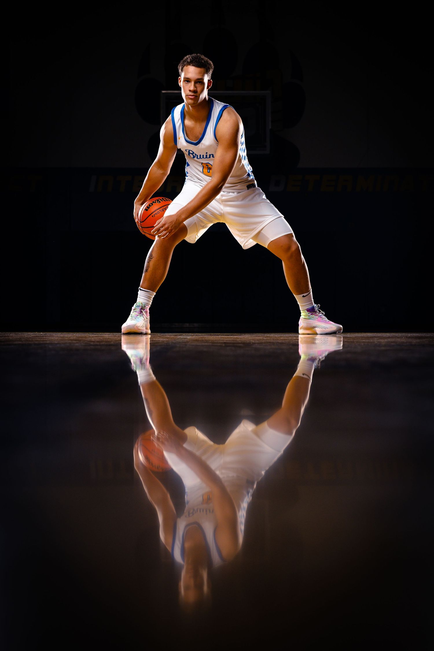 Basketball player in white jersey crouches in defensive stance on reflective court surface with dramatic lighting.