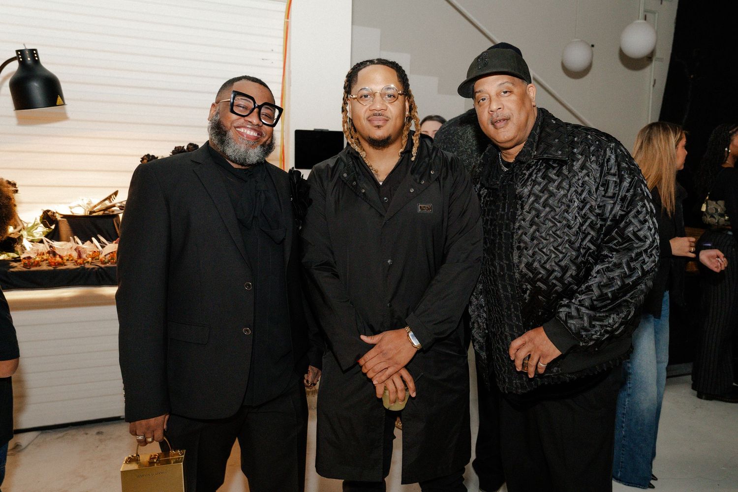 Three men pose together smiling at an indoor event, dressed in stylish dark outfits against a modern white interior.