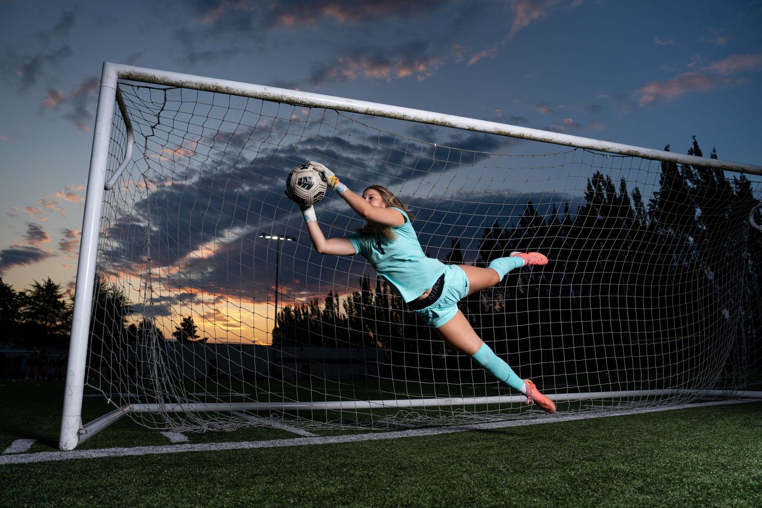Soccer goalkeeper in teal uniform makes diving save sequence against dramatic sunset sky.