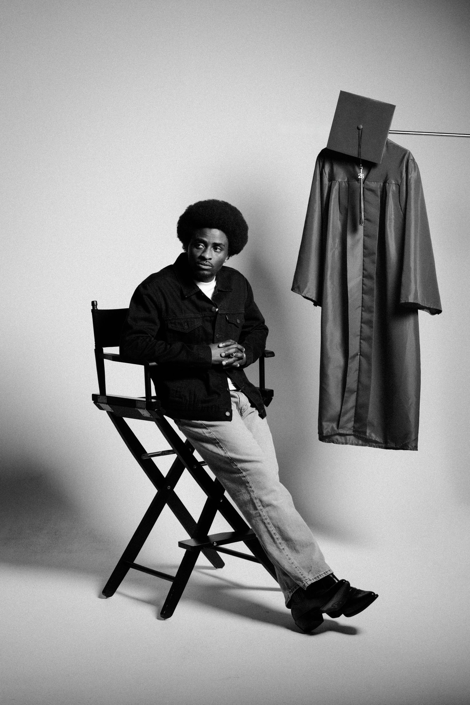 Young man with afro sits in director's chair beside hanging graduation cap and gown in black and white photo.