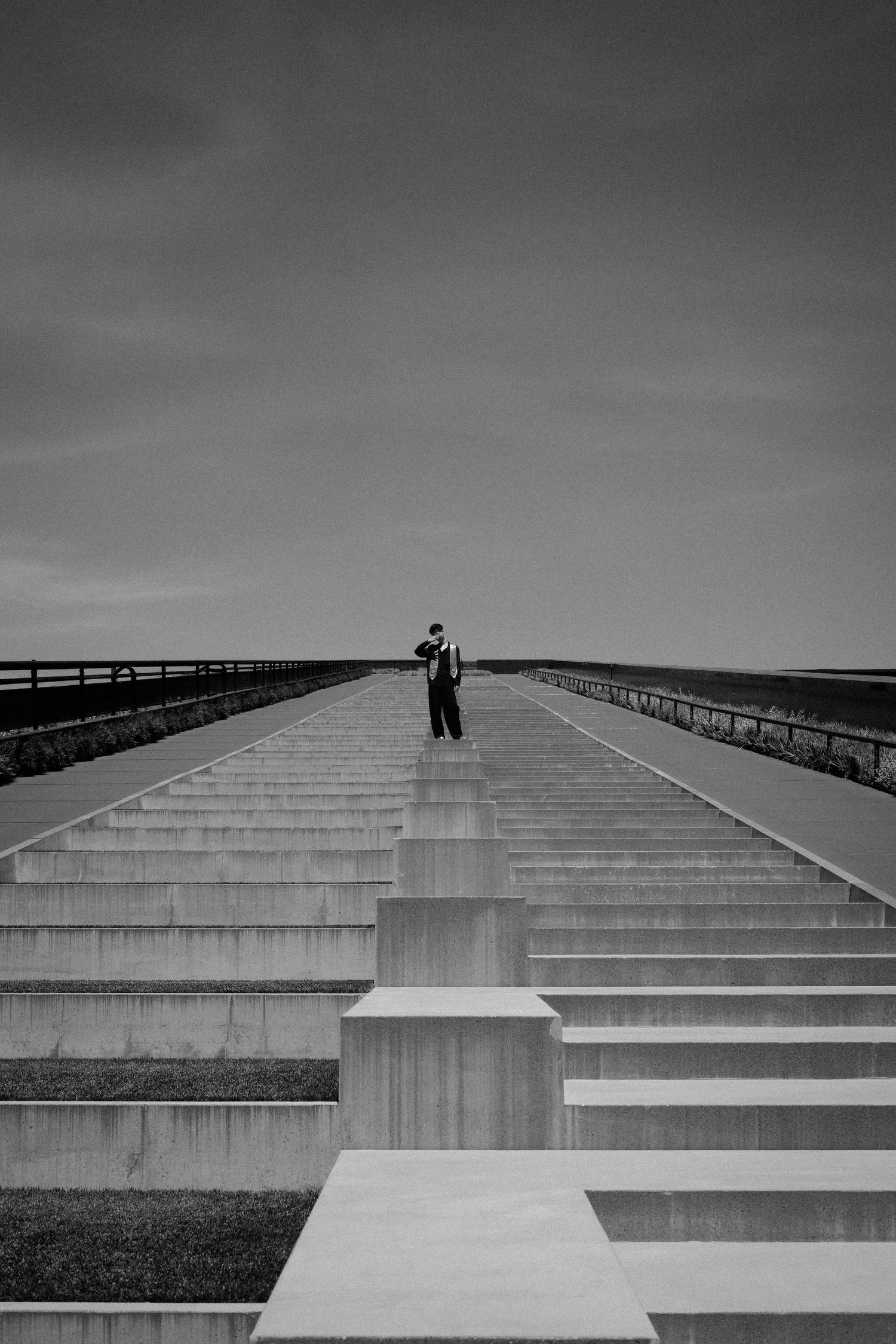 Black and white photo of a lone figure standing atop wide stone steps leading to a long bridge under cloudy skies.