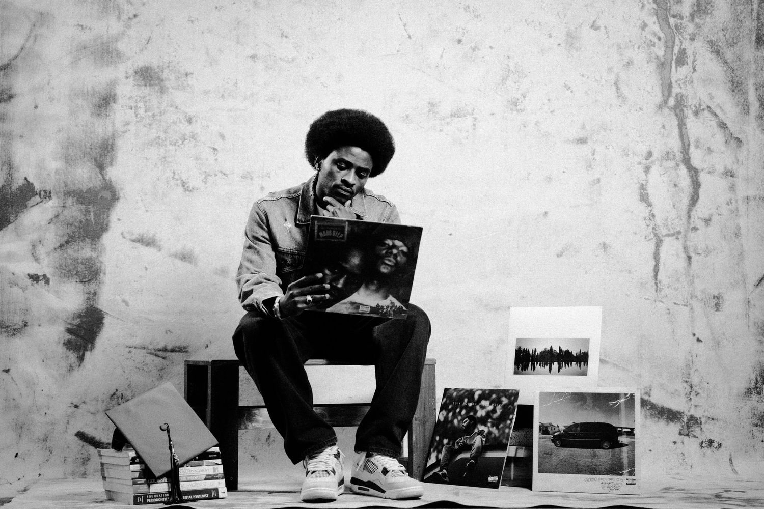 Black and white photo of a man with an afro sitting among vinyl records and photographs in a studio setting.