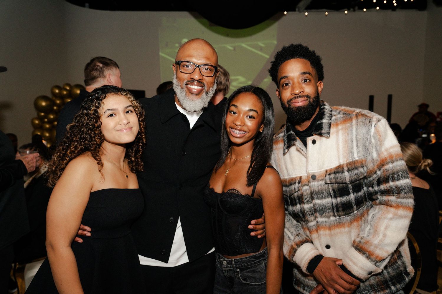 Four people smiling together at a festive event with gold balloons and green lighting in the background.