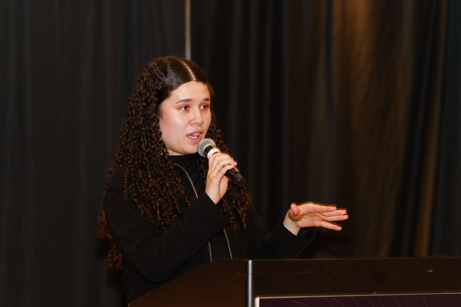 Young woman with curly hair speaking into a microphone at a podium during a conference presentation.