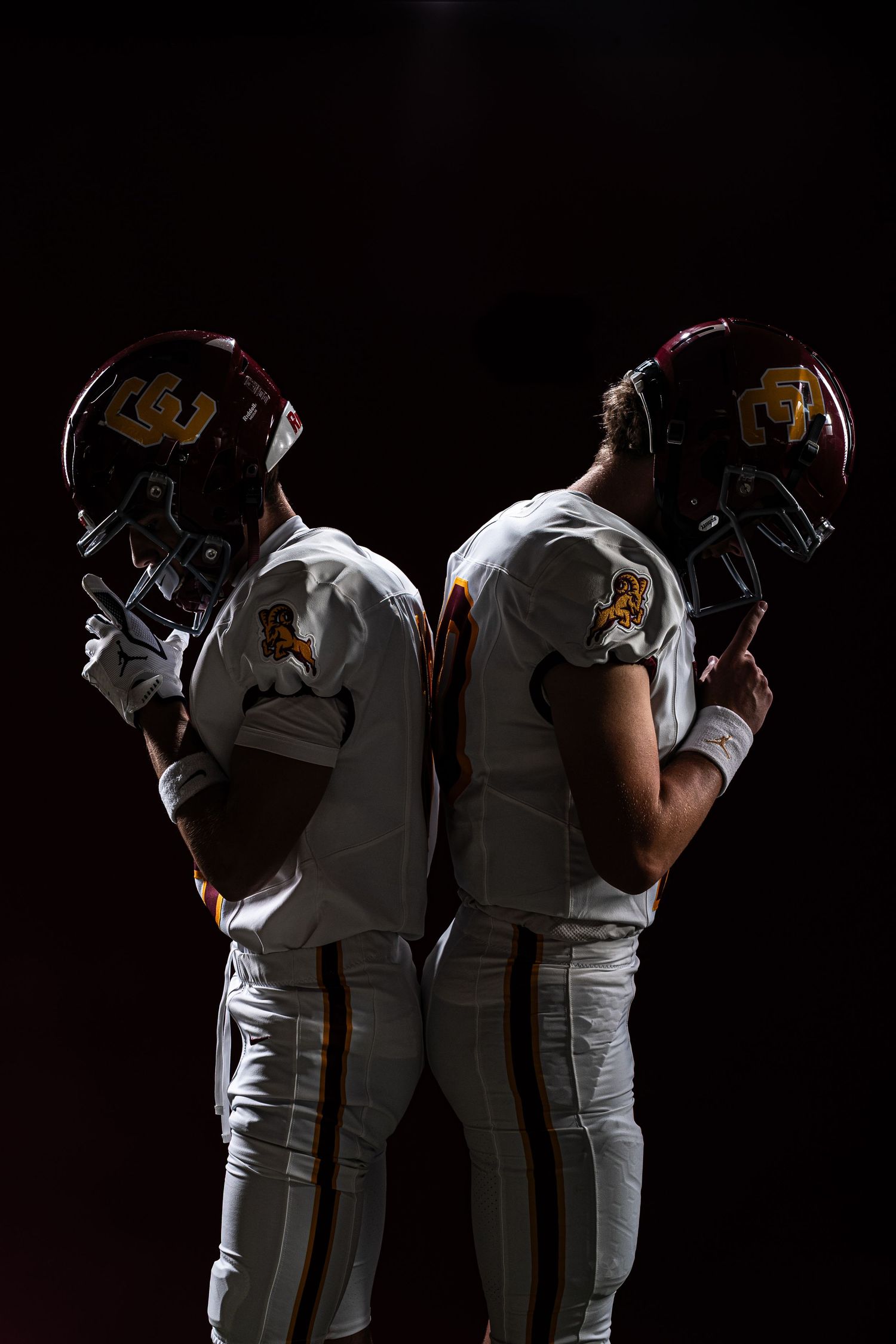 Two American football players in white and maroon uniforms stand back-to-back holding helmets against a dark background.